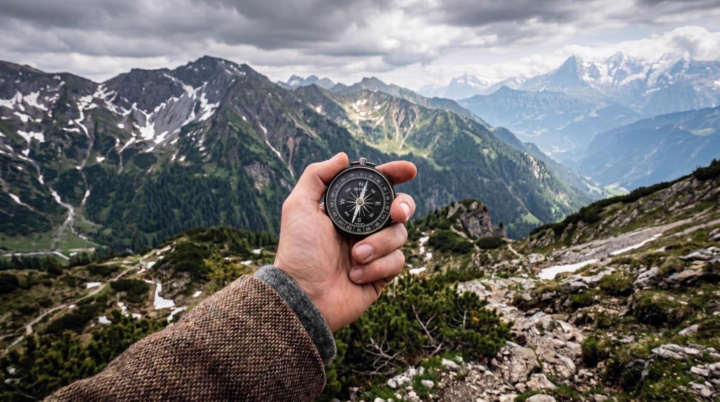 A hand holding a compass before a wide alpine panorama under dramatic clouds